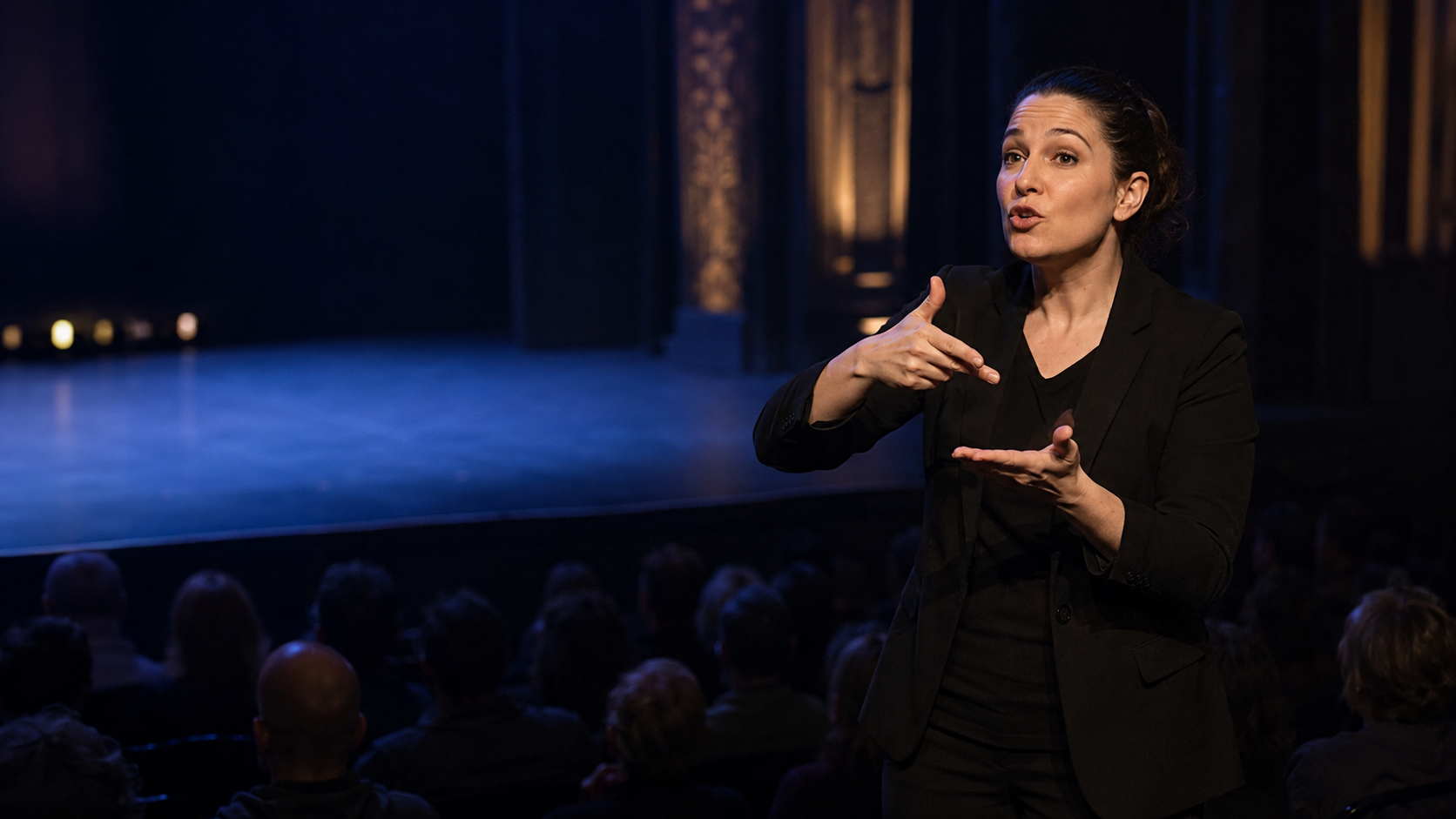 An ASL interpreter signing in a spotlight against a dark stage
